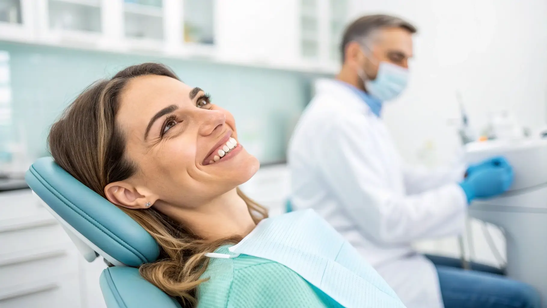 A smiling woman in a dental chair with a dentist in the background, soft focus. A professional and friendly patient smiling with a healthy smile
